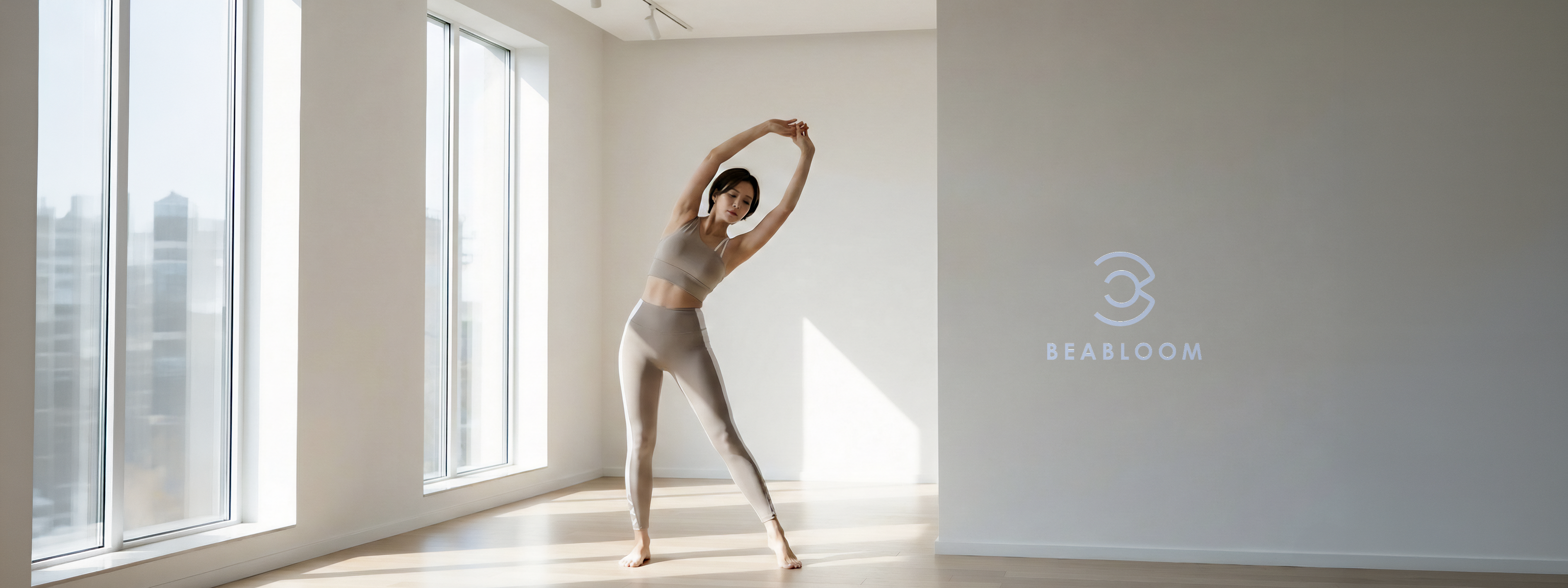 窓から差し込む朝の光の中、ベージュのスポーツブラとレギンスで体を伸ばす女性。BEABLOOMの着用イメージ