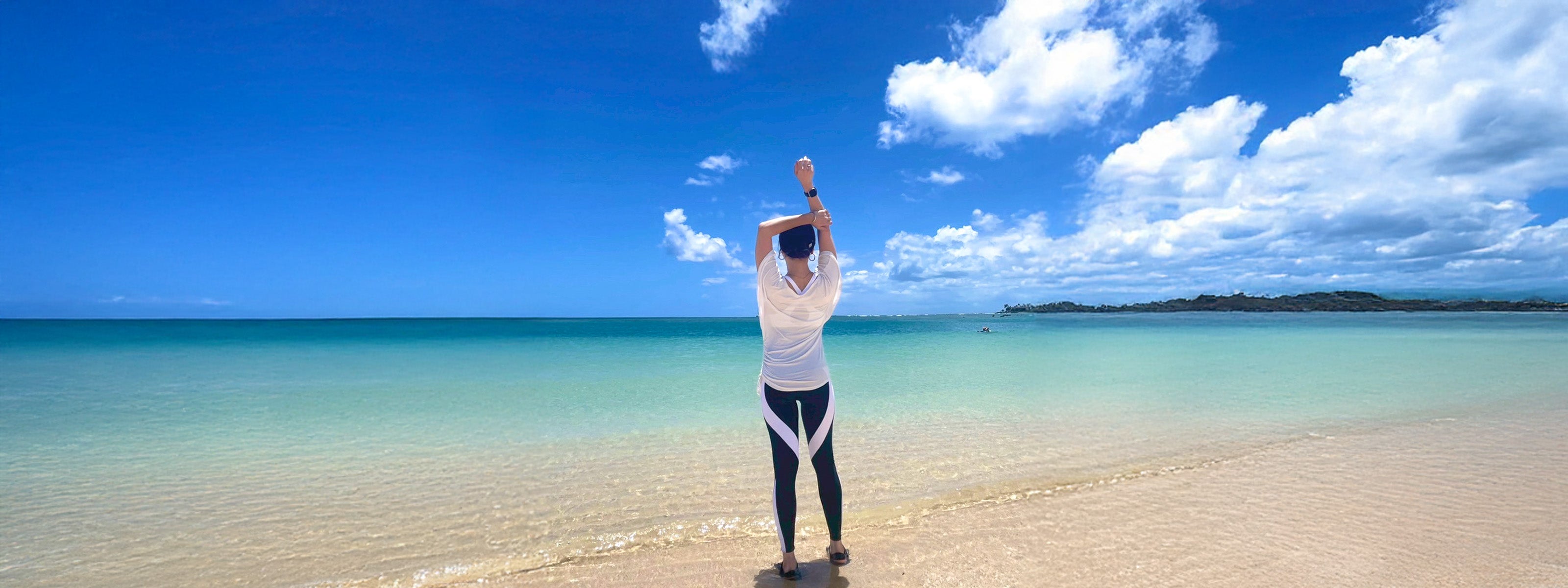 BEABLOOMのヨガウェア・ピラティスウェアを着用した女性が、ハワイの海岸でストレッチポーズをとる後ろ姿。鮮やかな青空とエメラルドグリーンの海が広がる自然の中で、心身をリフレッシュするシーン